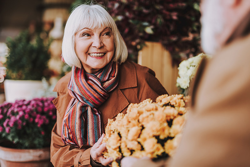 Very nice lady with white hair smiling outside at a table with yellow flowers in front of her
